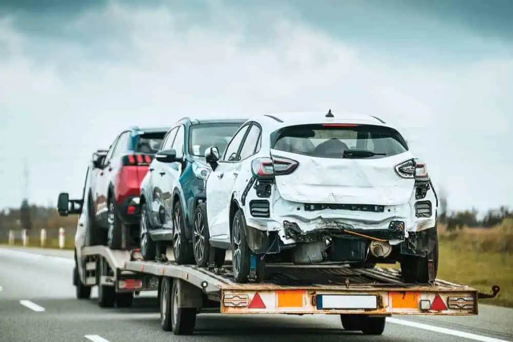 A tow truck transports two damaged cars, one white and one red, along a highway under a cloudy sky. Both vehicles display rear-end damage, with the white car being in the forefront on the flatbed. The background features a field and distant trees—a typical scene during 24/7 Towing in Queens County.