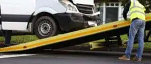 A white van with a damaged front bumper is being loaded onto a yellow tow truck ramp by a person wearing a yellow reflective vest. The scene appears to be taking place on the side of a road, showcasing the reliable roadside assistance in Queens County provided by Towing Services Queens County.