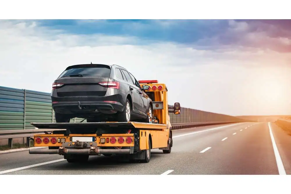 A black SUV is being transported on a yellow flatbed tow truck driving on a highway. The sky is partly cloudy, and the road is empty, lined with a guardrail and a sound barrier. This efficient roadside assistance in Queens County ensures vehicles are safely moved anytime.