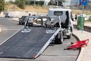 A flatbed tow truck with its ramp extended is parked on the side of a road. Nearby, two electric scooters lie on the ground, one red and one black, possibly prepared for towing. The background shows a mix of urban and natural elements—exemplifying 24/7 Towing Queens County services in action.