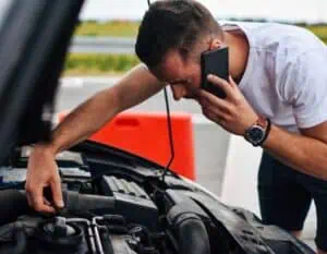 A man stands next to his car with the hood open, using his phone while inspecting the engine. Wearing a white t-shirt and a wristwatch, he looks intently at the engine components, possibly troubleshooting an issue. Bright orange traffic cones are visible in the background as he contemplates whether to call 24/7 Towing Queens County.