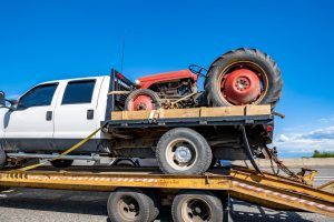 A white pickup truck from a renowned Towing Company in Queens County is seen towing a flatbed trailer carrying an old red tractor. The bright blue sky in the background and clear road indicate it is daytime. The tractor is secured on the trailer with yellow straps and wooden blocks.