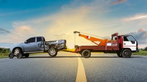 A tow truck with a red and white cab and yellow crane, offering 24/7 Towing in Queens County, is towing a severely damaged silver pickup truck on an empty road. The background features a sunset with a partly cloudy sky and green fields, making the serene landscape contrast with the day's unfortunate event.